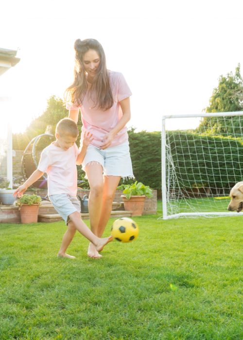 Young mother and son plays soccer with dog and have fun together. Happy family playing football with pet. Fun Playing Games in Backyard Lawn on Sunny Summer Day. Motherhood, childhood, togetherness.