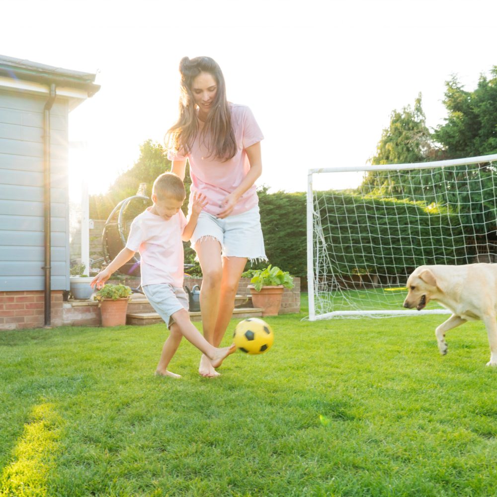 Young mother and son plays soccer with dog and have fun together. Happy family playing football with pet. Fun Playing Games in Backyard Lawn on Sunny Summer Day. Motherhood, childhood, togetherness.